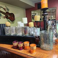 Photo showing multiple candle holders on a wooden surface with background featuring wall art, including clear glass holders and colored votive holders with metallic bands