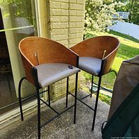 Pair of director style bar stools with tall black metal legs, round curved wooden backrests, and gray upholstered seats placed outside near a door.