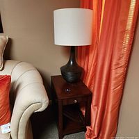 Wooden end table with drawer and slatted shelf beside a sofa with a decorative ceramic lamp on top.