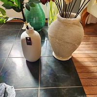 Two large ceramic vases placed on floor, one white minimalist design and one aged white with carved horizontal lines, featured with some decorative branches inside the aged vase.