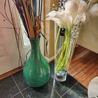 Two tall floor vases on black tile floor near a door and hardwood floor. One green painted woven-style vase and one clear glass cylindrical vase with faux flowers.