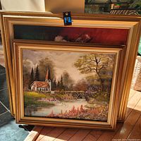 Photo showing two framed paintings stacked, with visible scene of church by pond in the forefront.