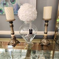 Full lot view of the two tall gray candle holders, large clear glass vase with gray stones inside, and white faux flower in the vase, arranged on a mirrored surface.