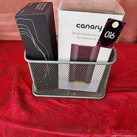 Photo showing the boxed Canary Smart Home Security System and another black box, all inside a gray metal mesh container.