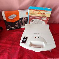 Photo showing Proctor Silex waffle maker with green and red indicator lights, plastic body, and the packaging for Micro Hot Plate and BBQ serving tray in the background.