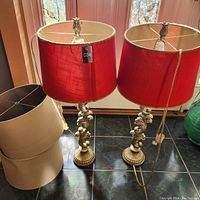 Two table lamps with red shades on chrome scroll bases with two off-white lampshades next to them on floor.