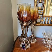 Three large amber glass and brass candle holders arranged on a wooden surface, under warm lighting. Shows the intricate brass bases and amber glass shades.