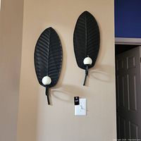 Front view of two black metal leaf-shaped wall candle holders mounted on a beige wall with white spherical candles placed on the holders.