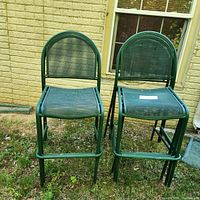 Two green outdoor metal mesh bar stools shown from the front, placed outside on grass with a brick wall background. Both stools have mesh metal seats and backs, with visible rust on the legs.