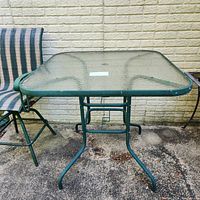 Square green metal framed outdoor bar table with glass top against a textured wall