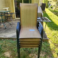 Front view of six stackable outdoor chairs showing metal black frame and light brown woven fabric seats and backs stacked one on top of the other outdoors on grass.