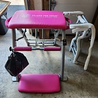 Full view of the Pilates Pro Chair showing pink padded seat, metal frame, and foam grip handles, with a black bag hanging on one side.