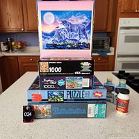 Photo of stacked puzzles and puzzle glue bottles on kitchen counter showing all items in lot and box art.