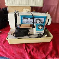 Side view of DressMaker sewing machine sitting inside bottom half of the carrying case, showing dials, stitch patterns, foot pedal and cords.