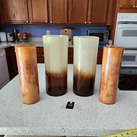 Four items including two tall glass candle holders with gradient chocolate latte coloring and two matching tall pillar candles with orange marbled design, arranged on a kitchen counter.