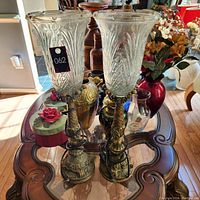 Pair of tall bronze candle lamps with decorative bases and large clear glass shades on a wooden table.