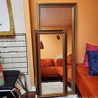 Two tall mirrors leaned against an orange wall near an orange couch, showing full height and frame details.