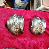 Two silver-toned decorative plastic wall sconces placed side by side on red fabric, showing overall shape and embossed detail.