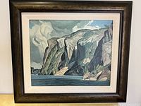 Framed glicee print showing a landscape of cliffs and water with cloudy sky. The frame is dark wood with slight damage but overall in fair condition.