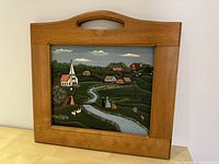 Frontal image of the Dutch landscape painting inside a wooden frame with a handle on top. The painting features a church, houses, river, people, and animals in a pastoral setting.
