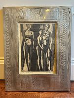 Framed print artwork of Gravür Baski by Oya Abaci showing five abstract figures in black and gray tones inside a textured patterned metal frame placed on wooden floor against white baseboard and wall.