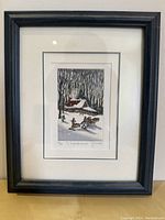 Photo of framed print showing a winter scene with a snow-covered cabin, figures skiing or sledding in snow, black wooden frame and white matting, excellent condition