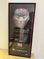 Photo of a framed poster featuring a detailed carved mask representing Tsimshian culture with event details below, mounted in a black frame with reddish matte.