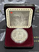 Photo of the display case interior showing the coin in a clear capsule and certificate card describing the coin's historical significance and specifications.