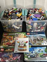 Two large transparent bins filled with assorted Lego bricks and pieces. Several Lego instruction manuals placed in front, representing Ninjago, Harry Potter, Marvel Super Heroes, and other themes.
