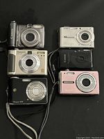 Top view showing all six digital cameras arranged in two columns of three, with Canon, Panasonic, GE, Casio, and Olympus brands visible.