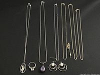 Full view of six silver jewelry pieces laid out: four necklaces including one with bird pendant, one with purple teardrop pendant, one pair of earrings with pearl accents, and one ring with clear stone.