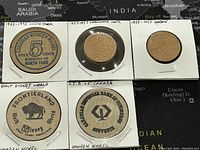 Front view of 5 wooden nickels in holders with handwritten notes labeling dates and origin including Canadian and London commemorative nickels, Walt Disney World Frontierland and Canadian Pike Company.