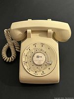 Front view of the almond-colored rotary dial telephone showing numbers and letters on the dial and coiled cord to handset.