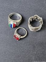 Top down view of three sterling silver rings showing multicolored stone ring, red gemstone ring, and textured band ring on dark background