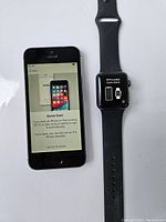 Photo of unlocked black iPhone showing Quick Start setup screen next to Apple Watch with black band showing pairing instructions.