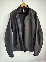 Full front view of men's Patagonia black softshell jacket hanging on wooden hanger. Front zipper partially open, zippered pockets visible. Jacket shows normal signs of use.