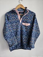 Front view of the blue Patagonia Synchilla pullover shirt with pink trim and geometric pattern hanging on wooden hanger.