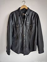 Full front view of vintage black leather Harley Davidson shirt on hanger, showing button-up front and chest pockets.