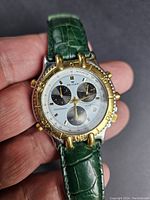 Close-up of the watch face showing silver dial, three black subdials, gold bezel with tachymeter, gold accents, and green leather strap in hand.