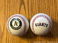 Photo showing two official baseballs, one with Oakland Athletics logo, the other with San Francisco Giants logo placed side by side on a wooden surface.