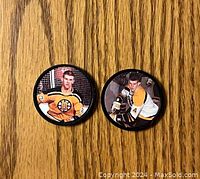 Two hockey coins with Bobby Orr images on top side, one in yellow Bruins jersey number 7, other in white and yellow jersey number 4, placed on wooden surface.