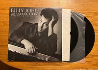 Front cover of Billy Joel Greatest Hits Volume 1 & 2 showing black and white photo of Billy Joel leaning on piano with vinyl partially pulled out.