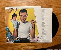 Front side of vinyl record sleeve featuring a man holding a puppet and printed lyrics, with the vinyl partially visible.