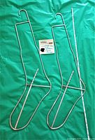 Pair of vintage metal wire sock stretchers or boot hangers laid flat on green fabric background with measuring tape beside one for scale.
