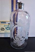 Front view of clear glass apothecary bottle with white etched floral design and metal stopper with cork.