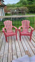 Pair of red resin Adirondack chairs on wooden deck with outdoor background.