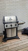 Full front view of Broil King Signet gas BBQ grill showing stainless steel body, three control knobs, side shelf, wheels, and propane tank storage doors.