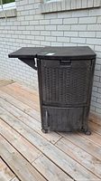 Brown resin cooler station with woven design, caster wheels visible, side shelf folded out. Positioned on wooden deck against a brick wall.