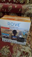 Photo showing front and side view of the Rove MyDash R2-4K dashcam box on patterned carpet with sofa background.