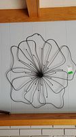 Front view of black metal flower wall art sculpture hung on a gray wall with wooden beam above. Sculpture features petal outlines made from curved wire and fine central spokes ending in small spheres.